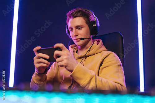 Foto Portrait of caucasian man pro gamer sitting on gaming chair, wearing headset, has live stream while playing mobile game on the smartphone at home
