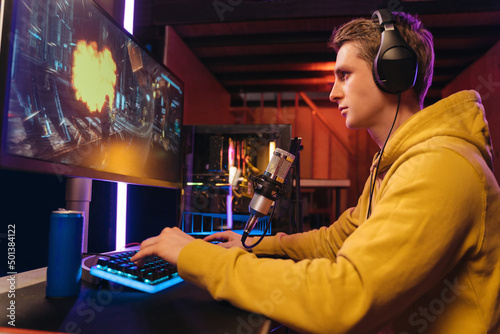 Concentrated professional gamer playing shooter video game on his powerful computer, streaming his participating in online cyber games tournament, drinking energy drink to focus on game late at night