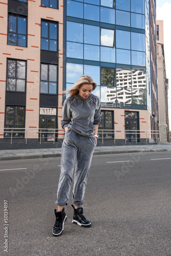 teen leisure. young blonde teen girl in sports gray velour costume stands casual with hands in pockets and looks down with cute smile on the street building background. lifestyle concept, free space