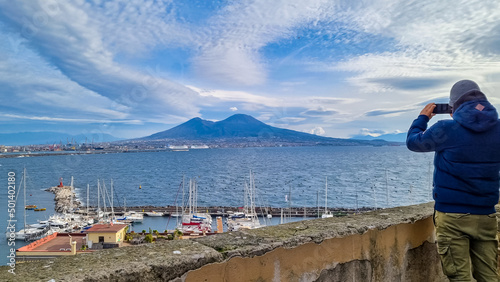 Fototapeta Naklejka Na Ścianę i Meble -  Tourist man taking picture with panoramic view from Castel dell Ovo in city of Naples, Campania, Italy, Europe. Bathing in the sun. Panoramic view on volcano mount Vesuvius. Ferries in harbor, Napoli