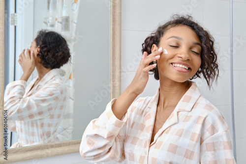 Happy african american woman applying moisturizer on her face near the mirror