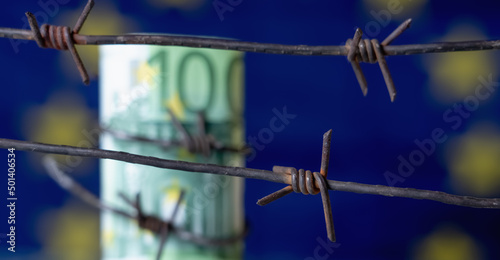 European Union currency wrapped in barbed wire against flag of EU as symbol of Economic warfare, sanctions and embargo busting. Selective focus on barbed wire