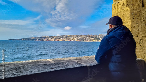 A tourist man enjoying the panoramic view from Castel dell Ovo (Egg Castle) on the city of Naples, Campania, Italy, Europe. Person leaning against the castle wall, bathing in the sun. Sea view