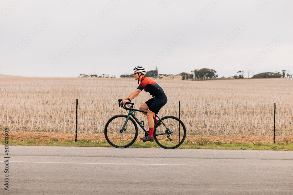 Obraz premium Side view of young woman cyclist in sportswear riding road bike against a field