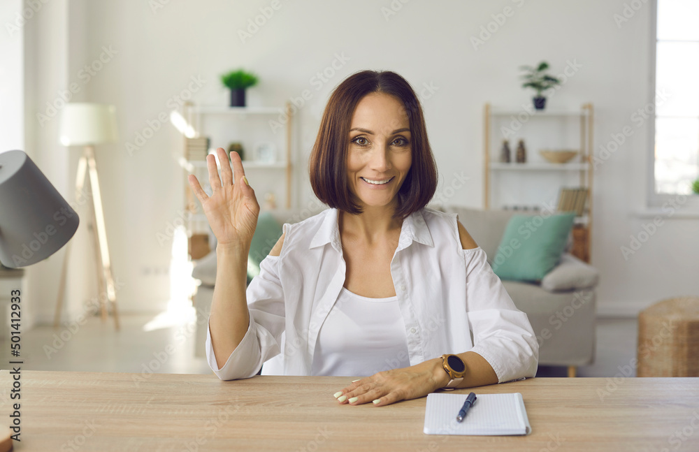 Happy beautiful woman sitting at work desk with notebook and pen at home, waving hello and smiling. Web cam laptop computer screen view of adult student in online lecture, videocall or videoconference Happy beautiful woman sitting at work desk with notebook and pen at home, waving hello and smiling. Web cam laptop computer screen view of adult student in online lecture, videocall or videoconference