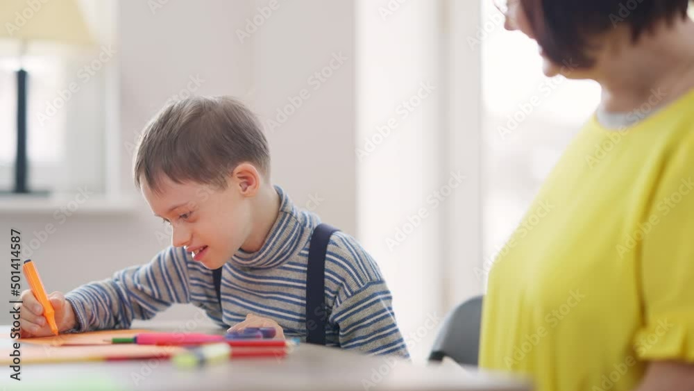 Autistic schoolboy painting talking with teacher in classroom in school. Portrait of excited curios Caucasian boy with mental illness studying indoors with blurred woman supporting child