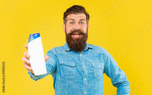 amazed brutal bearded man hold shampoo bottle on yellow background, haircare