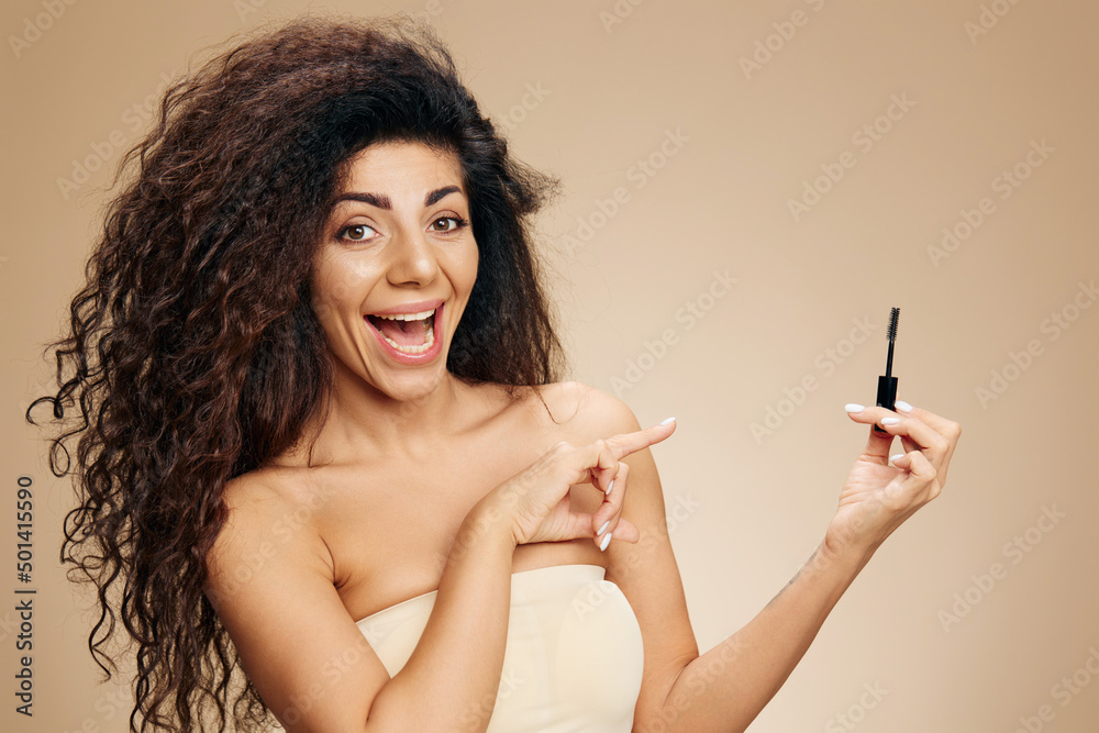 EYEBROW GEL AD CONCEPT. Excited happy tanned pretty curly Latin female point finger aside show mascara eyebrow gel brush cosmetic product posing isolated on pastel beige background. Copy space offer EYEBROW GEL AD CONCEPT. Excited happy tanned pretty curly Latin female point finger aside show mascara eyebrow gel brush cosmetic product posing isolated on pastel beige background. Copy space offer