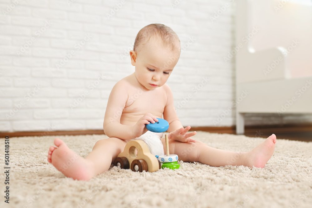 cute baby sits on a carpet in a disposable diaper plays with toys against a white brick wall. cute baby sits on a carpet in a disposable diaper plays with toys against a white brick wall.
