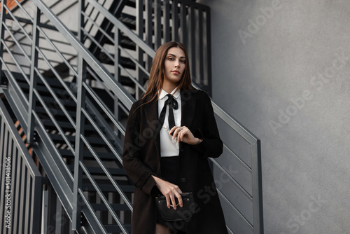 beautiful young business woman in black fancy suit with a black coat, white shirt and tie, and a black skirt with a purse walks in the city near a gray wall and metal staircase