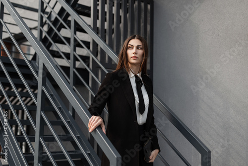 Beautiful professional business woman in a fancy black suit with a shirt, tie, and coat walks down the metal stairs outside in an office building