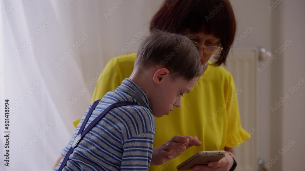 Side view focused autistic boy gaming on smartphone with woman helping child. Portrait of Caucasian concentrated curios son at home with caring mother. Support and individuality concept Side view focused autistic boy gaming on smartphone with woman helping child. Portrait of Caucasian concentrated curios son at home with caring mother. Support and individuality concept