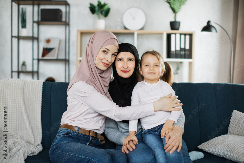 Beautiful islamic women generation: granny, mom and daughter are hugging, looking at camera and smiling while sitting on couch at home.