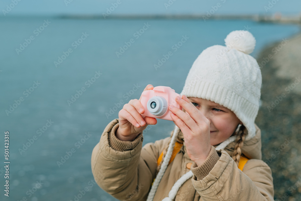 a little girl squinting photographs the autumn seascape on a childrens pink camera. the child is passionately engaged in his hobby: photography