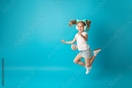 Fototapeta Naklejka Na Ścianę i Meble -  Funny young blonde girl in white t-shirt jumping isolated on blue background. People lifestyle concept. Listen music with headphones, sing song