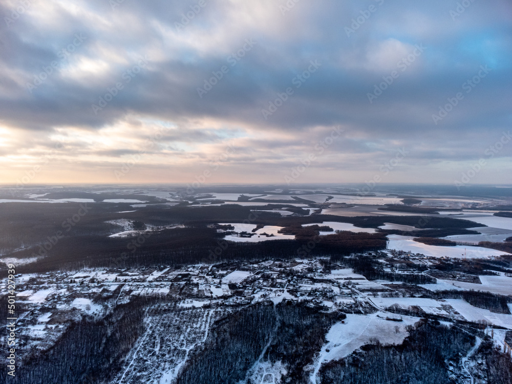 Obraz premium Winter aerial epic view on village, forest and fields covered in snow. Zmiyevsky region in Ukraine from drone. Sun shines through heavy sunset clouds