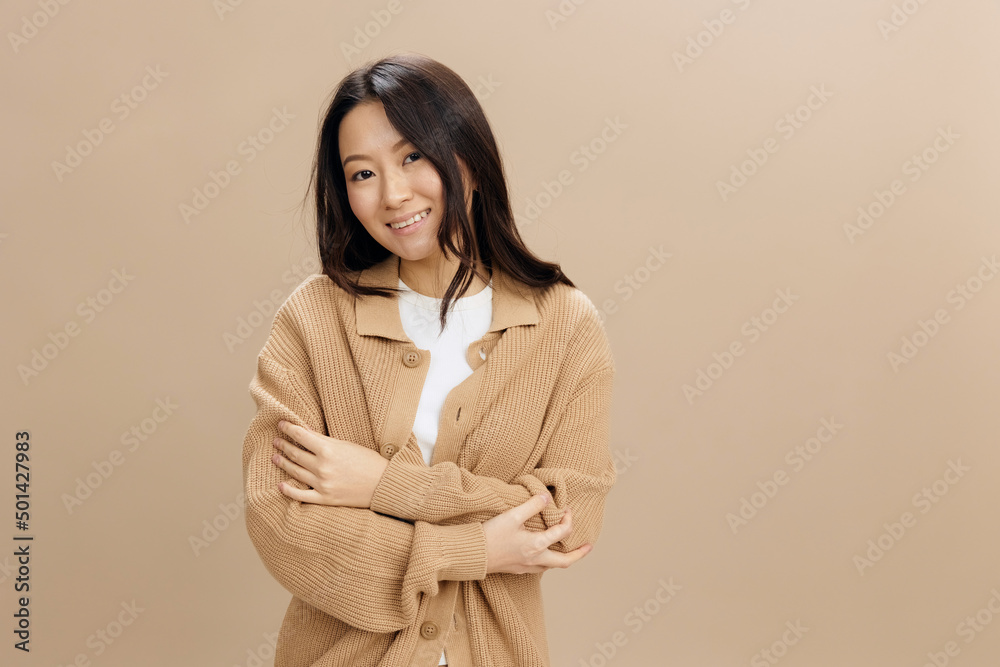 Cheerful happy smiling lovely Asian student young woman in warm sweater hugs herself posing isolated on over beige pastel studio background. Cool fashion offer. Seasonal Sale Fashion concept Cheerful happy smiling lovely Asian student young woman in warm sweater hugs herself posing isolated on over beige pastel studio background. Cool fashion offer. Seasonal Sale Fashion concept