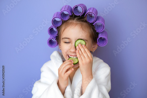 A funny little beautiful girl with curlers in her hair puts a piece of cucumber on her eyes and eats the second slice. Facial Skin Care, Moisturizing Natural Eye Mask. Loud children's laughter photo