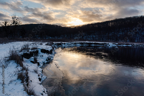 Winter view on frozen river curve with scenic sun reflection. Zmiyevsky region on Siverskyi Donets River covered in snow in Ukraine. Sunset sun in clouds above woody hill