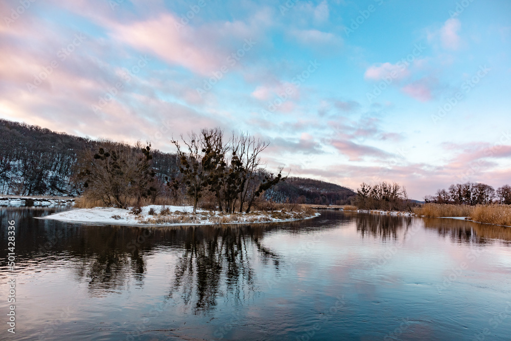 Winter river with scenic mirror reflection of bare trees on snowy coast and vivid evening cloudy sky. Zmiyevsky region on Siverskyi Donets River in Ukraine