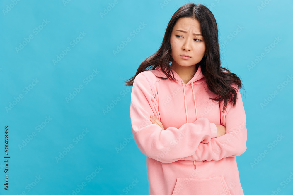 Fototapeta premium Upset sad cute Asian student young lady in pink hoodie sweatshirt folds hands posing isolated on over blue studio background. The best offer for ad. People Emotions for Everyday concept