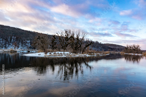 Winter river with scenic mirror reflection of bare trees on snowy coast and scenic vivid evening cloudy sky. Zmiyevsky region on Siverskyi Donets River in Ukraine © Kathrine Andi