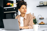 Perplexed african american curly-haired pretty girl, student or company worker working remotely from home, using laptop, looking away worriedly, thinking about project, deadlines