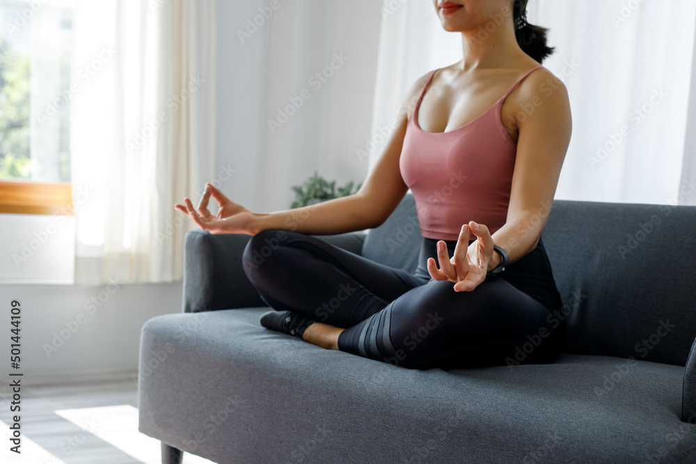 Yoga, gym, fit, sporty, Healthy, lifestyle, sport, and recreation concept. Peaceful millennial Asian woman doing yoga in lotus pose on the sofa,