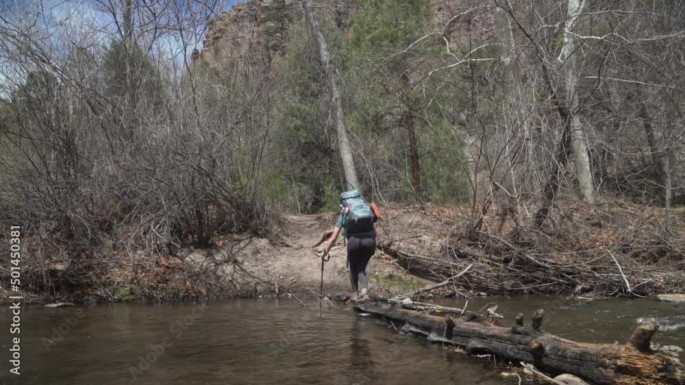 Female backpacker carefully crosses stream on log while hiking in New Mexico