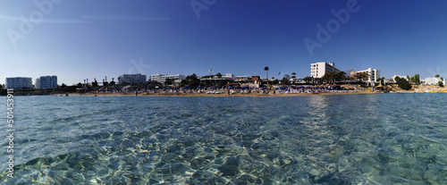 Protaras. Famagusta area. Cyprus. Panorama of Fig Tree Bay beach, people sunbathing and swimming, hotel buildings behind the beach against the sky. View from the sea.