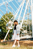Lonely young hipster woman walking at festival on background of ferris wheel and looking down. Smiling curly caucasian woman with hat in amusement park, lifestyle