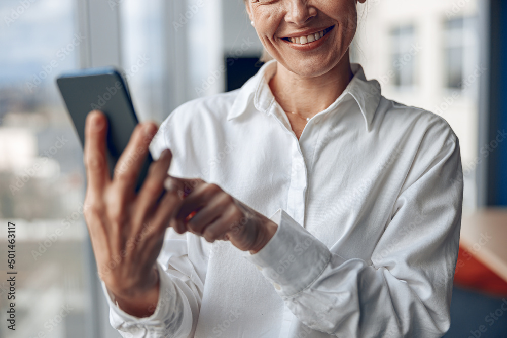 Smiling woman standing in office. Businesswoman with mobile phone in hand looking at smartphone. Smiling woman standing in office. Businesswoman with mobile phone in hand looking at smartphone.