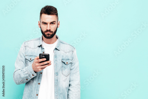 Handsome confident model.Sexy stylish man dressed in jacket and jeans. Fashion hipster male posing near blue wall in studio. Holding smartphone. Looking at cellphone screen. Using apps © halayalex