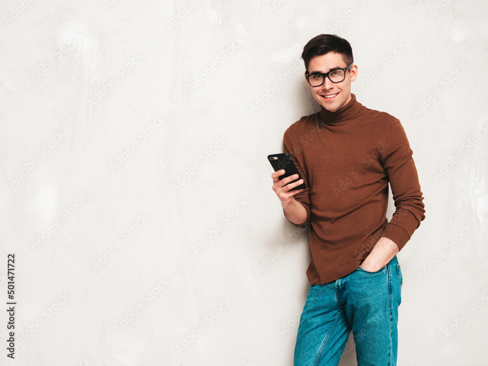 Handsome smiling model. Sexy stylish man dressed in brown turtleneck sweater and jeans. Fashion hipster male posing near grey wall in studio.Holding smartphone. Looking at cellphone screen. Using apps Handsome smiling model. Sexy stylish man dressed in brown turtleneck sweater and jeans. Fashion hipster male posing near grey wall in studio.Holding smartphone. Looking at cellphone screen. Using apps