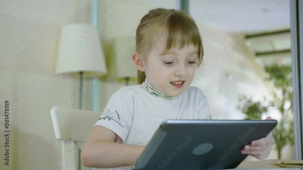 Funny Curious Child Little Girl Using Digital Tablet Technology Device Sitting At The Table Singing a Song Listening To Music Surfing Internet Play Game At Home Funny Curious Child Little Girl Using Digital Tablet Technology Device Sitting At The Table Singing a Song Listening To Music Surfing Internet Play Game At Home