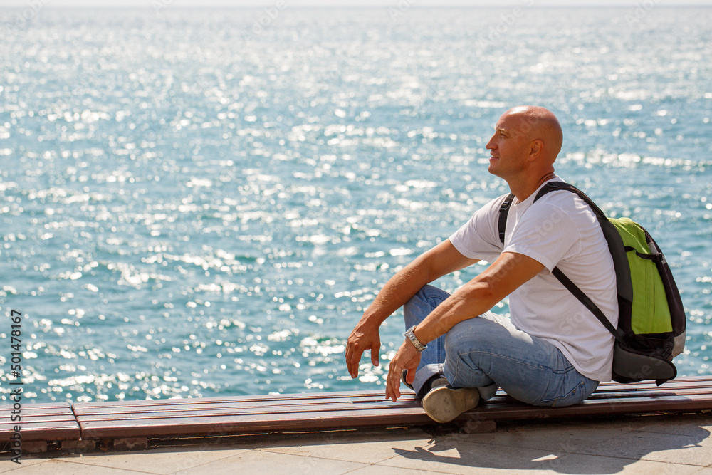 Portrait bald man on background sea. Tourist guy sitting on embankment near the pier enjoys a sunny day in casual clothes with a backpack looks around