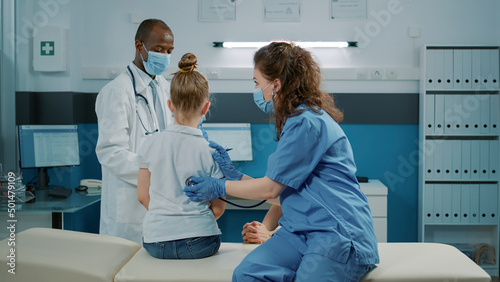 Medical assistant consulting child with stethoscope in cabinet at checkup appointment. Nurse using tool to measure heartbeat and pulse  giving assistance and support to kid and mother