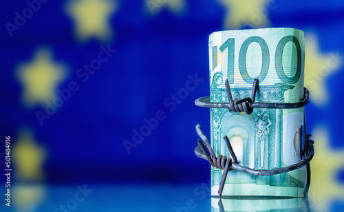 European Union currency wrapped in barbed wire against flag of EU as symbol of Economic warfare, sanctions and embargo busting. Horisontal image. Copy space. photo