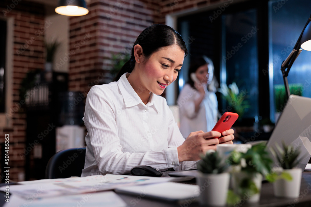 Happy smiling asian office worker sitting in office workspace while texting somebody on modern smartphone device. Businesswoman at work sending messages on social media platform using mobile phone.