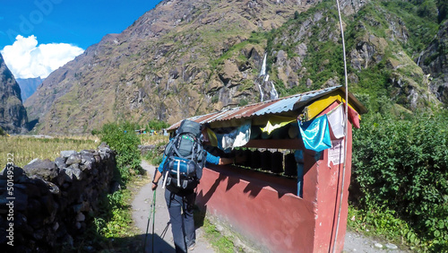 A man with big hiking backpack spinning the prayer wheels in a small Himalayan Village, Tal, along the Annapurna Circuit Trek. In the back there are high mountain chains overgrown with greenery.