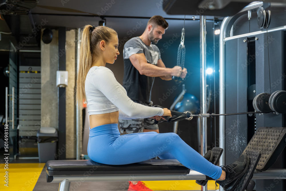 Obraz premium Sporty people working out in the gym. Attractive blonde hair young woman using cable crossover machine. Handsome man pushing down cable cross machine at health club.
