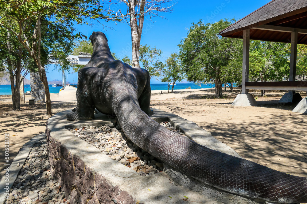 Fototapeta premium Backside view on a gigantic, venomous Komodo Dragon roaming free in Komodo National Park, Indonesia. The dragon is looking at the beach. Dangerous animal in natural habitat. Small pavilion on the side