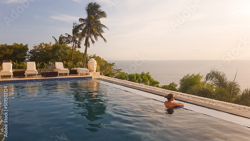A young woman leaning at the edge of an infinity pool and looking at the sun setting into the sea. There is a big palm tree in front. Calmness and serenity. Luxury hotel in Lombok, Indonesia.