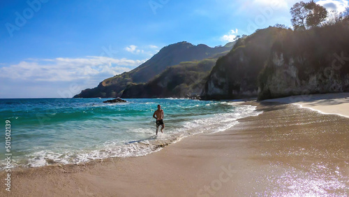 A man running along the shore, chasing the waves on idyllic Koka Beach. Hidden gem of Flores, Indonesia. He is having a lot of fun, collecting happy moments. Adventure and discovering while travelling photo