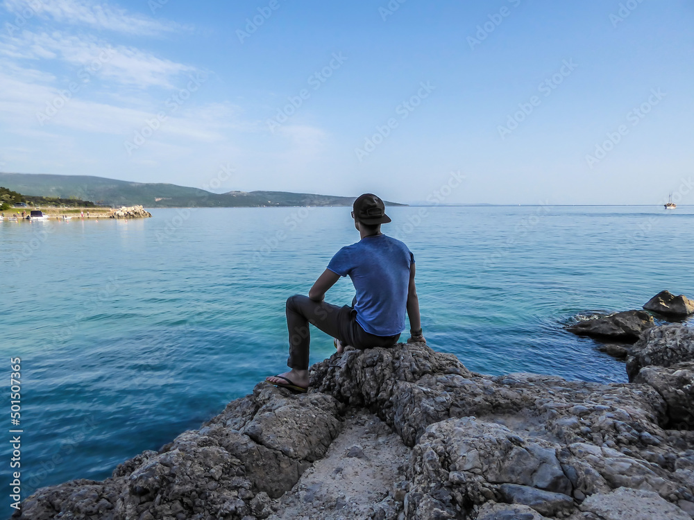 Man is a full cap sits at the stony shore of the Mediterranean Sea and looks at the calm surface of the water. He is calms and peaceful. Water has many shades of blue. In the back there are islands