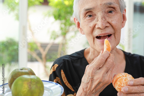 Happy healthy senior woman eating tangerine deliciously,asian old elderly opening her mouth wide to taste delicious organic orange fruit in the morning,nutrition,healthy lifestyle,health care concept