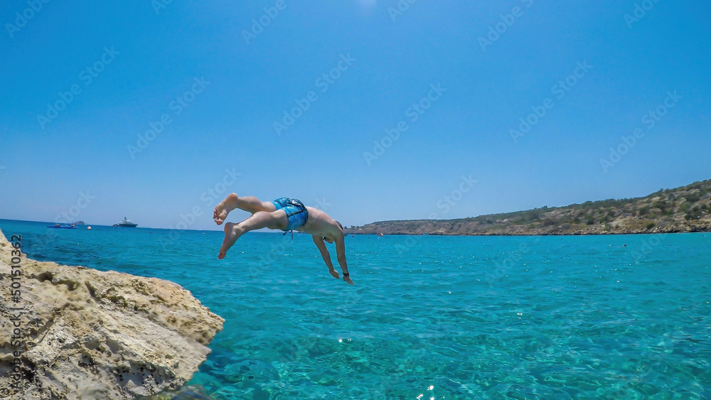 A young athletic-build man jumping into the sea in Cape Greco, Cyprus. Water is crystal clear, shimmering with many shades of blue. Jump of joy. Man is wearing blue swimsuit. A young athletic-build man jumping into the sea in Cape Greco, Cyprus. Water is crystal clear, shimmering with many shades of blue. Jump of joy. Man is wearing blue swimsuit.