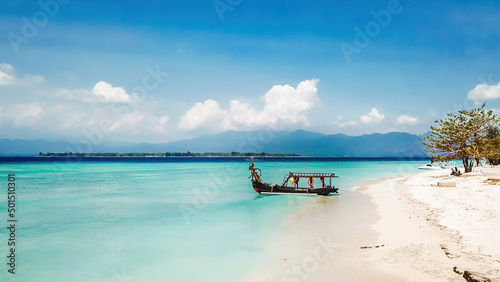 A boat parked next to the shore on Gili Air, Lombok, Indonesia. Beautiful and clear water. In the back visible Mount Rinjani. Some trees on the shore, few clouds on the sky.