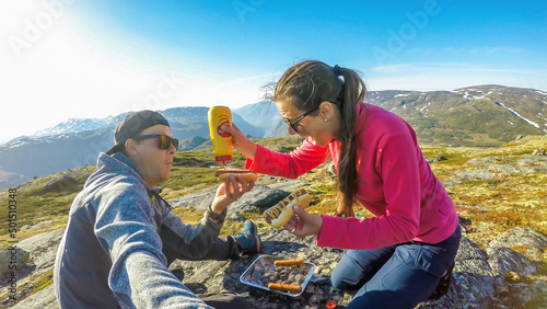 A couple having a grill in the tall mountains. Few sausages are being grilled. Couple uses portable one time grill, picnic grill. Girl spreads the mustard over the hot dogs. Camping in wilderness. © Chris