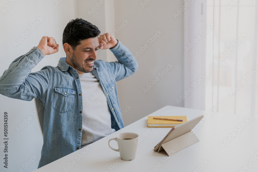 handsome young man smiling with arms outstretched while looking at a tablet sitting at his desk